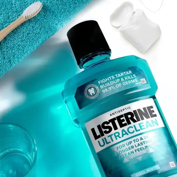 Listerine Ultraclean Cool Mint laid flat on a sink surface. A pack of floss, a toothbrush, a washcloth and a cup also laying flat surround the Listerine bottle