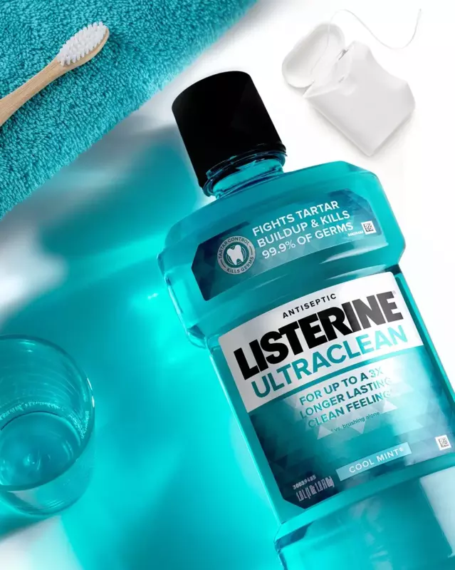 Listerine Ultraclean Cool Mint laid flat on a sink surface. A pack of floss, a toothbrush, a washcloth and a cup also laying flat surround the Listerine bottle.