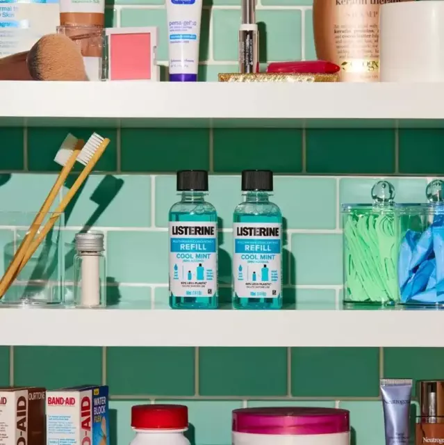Three white shelves are shown and behind is a teal tile wall. Several toiletries are shown such as lotions and toothbrushes but the image focuses on two Cool Mint Zero refill bottles.